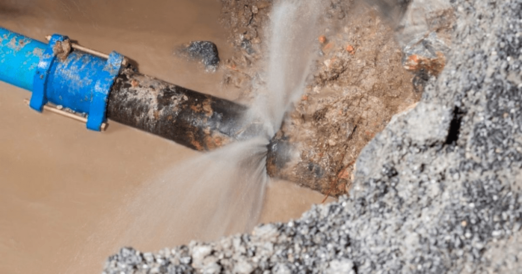 A képen egy sérült vízcső látható, amelyből víz szivárog egy repedésen vagy lyukon keresztül. A cső részben a talajba van ágyazva, és a sérült szakaszból erőteljesen spriccel ki a víz. A környező terület nedves és sáros, ami arra utal, hogy a szivárgás már egy ideje fennáll. Ez a kép szemlélteti az infrastruktúra-karbantartás fontosságát és azt, hogy a szivárgó csövek milyen károkat és vízpazarlást okozhatnak.